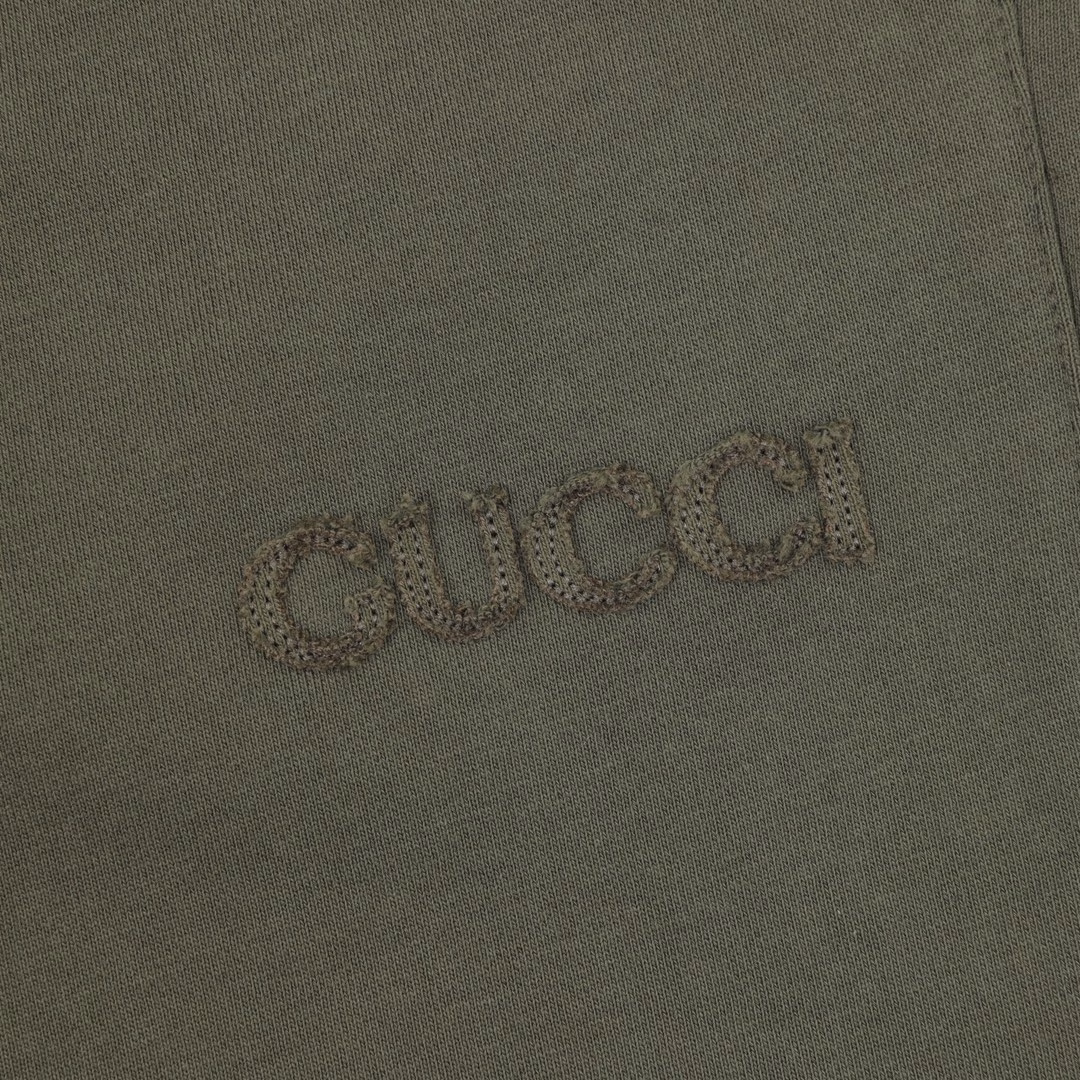 GUC letter embroidered shortsSingle-sided fish scale fabric woven with cotton as the base, the front is a smooth knitted surface, and the skin-touch surface is formed into a three-dimensional terry loop through the floating thread knitting technology of the knitting machine, which increases the fluffiness and delicate touch. The terry loop structure absorbs a lot of water and evaporates quickly, and it is not easy to loosen and deform after multiple washings. The same color fabric is cut into the brand letter pattern by laser, and the patch is temporarily fixed on the base fabric through hot melt adhesive film to ensure that the position is completely consistent with the design drawing. The same color thread is embroidered along the inner contour of the patch through a flat embroidery machine to form an invisible fixing layer to prevent the patch from shifting. By actively intervening in the shrinkage potential of cotton fibers, the fabric has achieved a leap from changeable to stable, improving dimensional stability, and is not easy to deform and wrinkle.